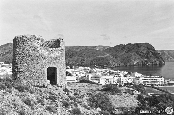 An Abandoned Windmill overlooks Las Negras surrounded by hills, with white buildings lining the shore and the sea visible in the background. The image is in black and white, highlighting the contrast between the ruins and modern architecture.