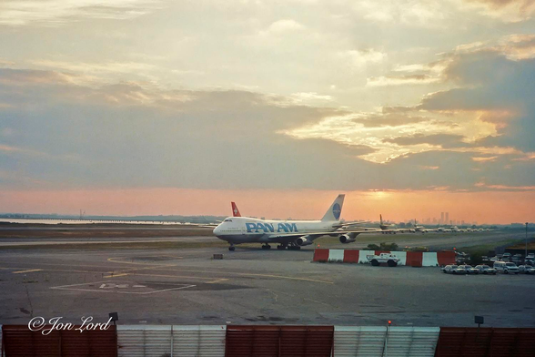 This is a scanned colour photo of an sunset scene at JFK airport with a Pan Am Boeing 747, a queue of aircraft waiting to takeoff and the Manhattan skyline in the background. New York (1989).

Stretching across the base of the image from left to right is a blast fence (like a Venetian Blind except about 3 or 4 metres high) the colours alternating from orange to white every 5m. The ground extends upwards to the 1/3 up level with the evening sky above. In the middle ground and again stretching from left to right is an expanse of concrete with some cars parked on the far right side. In the centre of the image is a Pan Am Boeing 747, taxiing from right to left, viewed obliquely with the front and left side in view and at a distance of about 100m. The fuselage has a grey underside and brilliant white above with 'Pan Am' in large pale blue lettering. On the white tail is a pale blue circle with Pan Am in the centre. On the far side of the 747 is a parallel taxiway with a long queue, about 1km, of aircraft facing away from the camera and waiting to takeoff on the runway that's parallel and beside the queuing aircraft. Beyond the runway is a narrow strip of water with a long and low bridge extending over the bay. On the horizon between the centre of the image and right side is a silhouetted view of the distant, southern New York City skyline with the former World Trade Centre twin towers visible. Above the city and stretching across the photo is a golden sunset and partly cloudy sky.