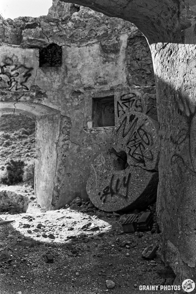 A monochrome image inside the ruins of an abandoned windmill with crumbling walls, featuring graffiti and a large, weathered mill-stone wheel on the ground. Sunlight filters through, illuminating the rugged textures and remnants of the site.