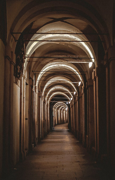 This is a photograph of a dimly lit, narrow, arched walkway with stone columns and walls. The ceiling has repeating, ribbed arches lit up by warm, white lights. The floor is made of aged, brown tiles.