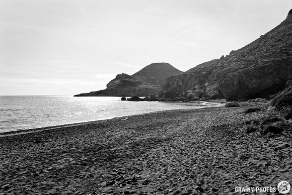 A serene black and white beach scene featuring gentle waves lapping at the shore, surrounded by rocky cliffs and distant hills under a soft sky, conveying a sense of tranquility and natural beauty.