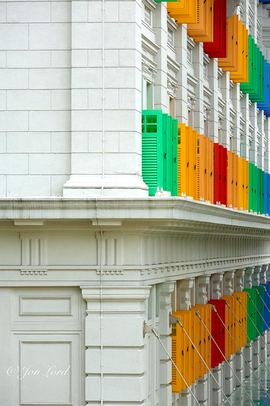 This is a colour photo in portrait format of the Old Hill Street Police Station, taken from the corner and looking along the front facade. Singapore (2013).

The view of the building is oblique with the left side facing the camera and the front stretching away into the distance from the corner to the right edge of the photo. Extending vertically through the centre of the photo is the corner of the building. From the corner and extending to the left margin is a white masonry wall, almost a faux marble in appearance, with a ledge reaching outwards about one metre from the facade. Above is a mock brick pattern with large rectangular shapes making up the bricks. Below is a little more elaborate with a short column like pattern below the ledge and a frame like adornment on the building's side. The front of the building is far, far grander, with the stonework in the same white colour, tall square pillars between the large rectangular windows that are almost hidden from view. In view are three floors. Each floor has multiple, brightly coloured, open shutters in front of their windows: set in an almost rainbow order. On the lower floor the shutters start with about six yellow shutters, then three red, six yellow, three light green, three dark green, then six blue before disappearing out of view. The floors above have a similar, but slightly differing order.

The building has a total of 927 windows, was a police station and is now the Ministry of Communications and Information.