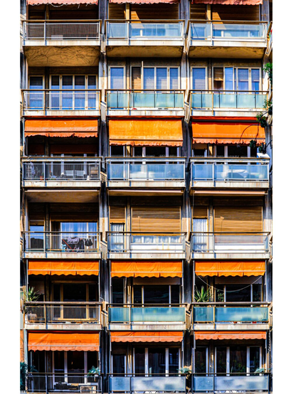 This photo is of a close-up view of an apartment building’s facade with several rows of balconies. The architecture is weathered concrete and a mix of metal and glass railings. There are bright orange awnings which are visible on multiple levels. They contrast against the neutral tones of the structure. Some of the windows are partially obscured by wooden shutters or curtains and a few balconies contain small plants and items like drying racks.