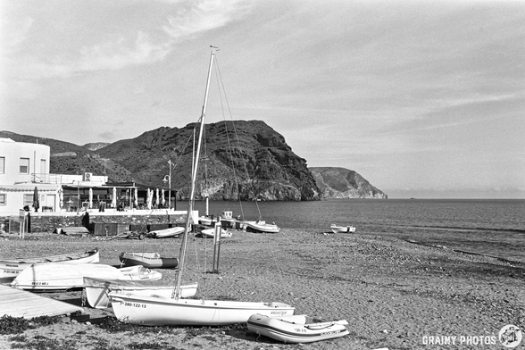 A serene black-and-white coastal scene featuring a sandy beach with small boats, a sailboat on the shore, a nearby restaurant, and a backdrop of a mountainous landscape under a clear sky.
