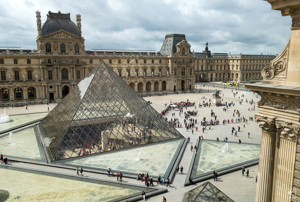 Color photo from a window of the Louvre museum in Paris looking down on the glass pyramid entrance situated in the middle of a large plaza - the Napoleon Courtyard - between the museum’s building wings. Surrounding the glass pyramid on three sides are shallow triangular pools with fountains. On the entrance side of the pyramid are lines of people queueing to get in and you can see people within the pyramid.