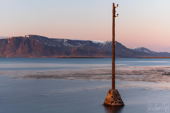 A single pole on a small platform stands in the water with a mountain range in the background during twilight