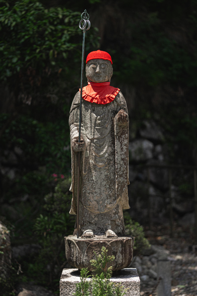 this is a photo of a Jizō statue wearing a little red hat and collar. I took this photo on a hike at Minoo falls, close to Osaka. specifically this statue was part of temple grounds which we found close to the beginning of the trail. it was a sunny day. the statue is surrounded by greenery. it was very quiet. us tourists were probably the loudest thing in the area. well I saw that in some of the faces of the local people. I had to remind myself to be a little calmer again, but it's hard not to get excited sometimes. 