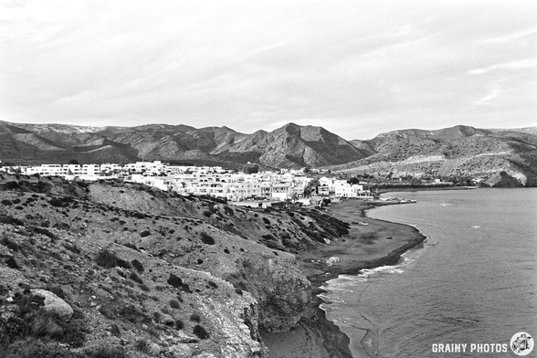 Black and white landscape of Las Negras nestled between mountains, featuring white buildings along a sandy shore and tranquil waters. The scene captures the natural beauty and serene atmosphere of the Mediterranean coastline.