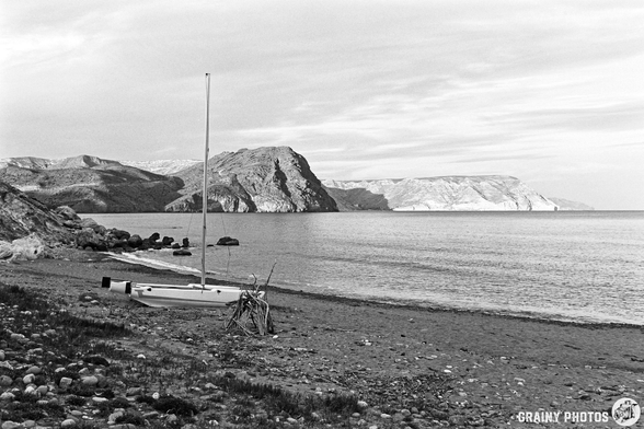 A serene black-and-white landscape featuring a small sailboat on a rocky beach, with gentle waves lapping at the shore. In the background, dramatic cliffs rise above the calm waters under a cloudy sky.