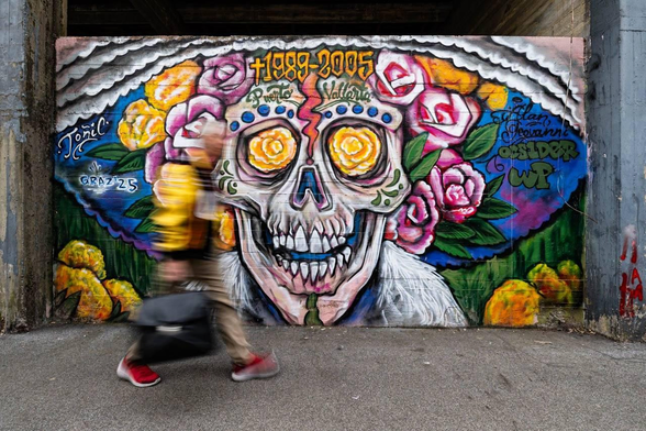 Ein farbenfrohes Mural eines mexikanischen Totenschädels an einer Betonwand wird von einer vorbeihuschenden Person in Bewegungsunschärfe kontrastiert.