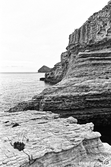 A monochrome image of rugged cliffs rising above the calm sea, with layered rock formations. In the distance, a small island is visible against a cloudy sky. The scene conveys a sense of solitude and natural beauty.
