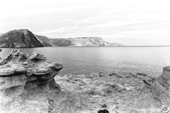 A black and white coastal landscape featuring rugged rock formations in the foreground, with calm waters reflecting a distant cliffside. The sky is overcast, enhancing the serene and dramatic atmosphere of the scene.