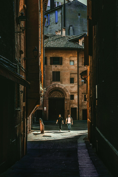 This potograph of a narrow, sunlit alleyway in Bologna (Via De' Pepoli). Two people, one in a dark t-shirt and a one in a white t-shirt, walk towards the camera. The alley is flanked by tall, old, brown/orange brick buildings with wooden doors and windows. Black lanterns hang on the left wall. The cobblestone ground reflects the warm, late afternoon light.