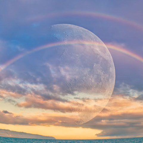 A large moon in superimposed on an image of two rainbows over the ocean. rain clouds and mountains in the background and a yellow glow from the sunrise.