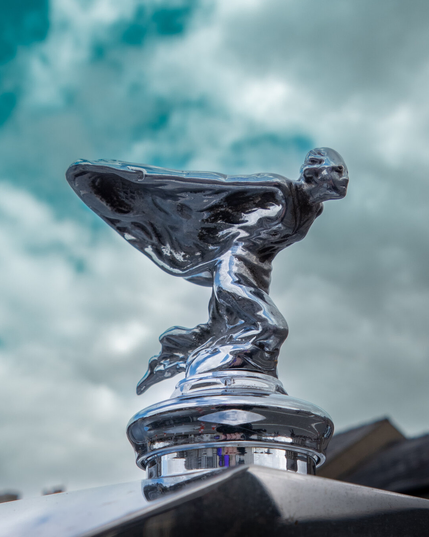 A close-up, low-angle shot of the iconic Rolls-Royce 'Spirit of Ecstasy' hood ornament featuring a woman leaning forward with her arms outstretched and robes billowing behind her like fairy wings. The chrome statuette reflects the light under a bright sky with soft white and blue clouds.