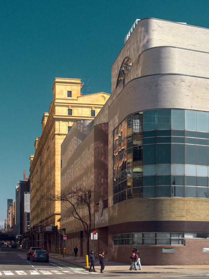 A building with about 4 floors with a rounded corner and glass wall reflecting another building across the street. There are several people crossing the street.