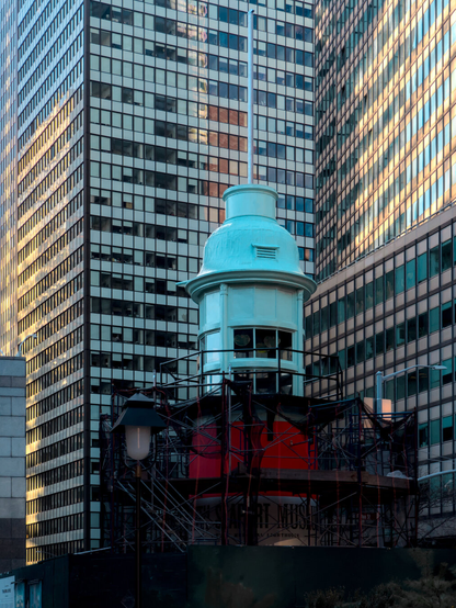 The teal Titanic memorial lighthouse beneath modern skyscrapers. The bottom of the lighthouse is red and under renovation.