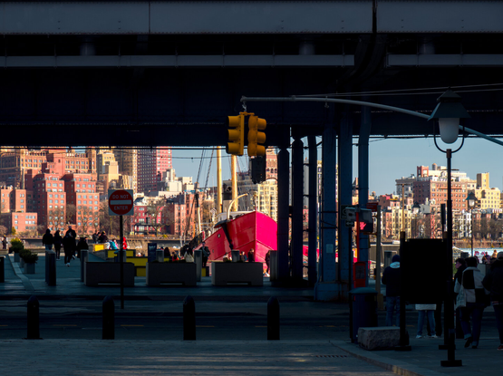 Looking from beneath a freeway viaduct at a red vintage ship and buildings across the East River in Brooklyn in the golden sun.