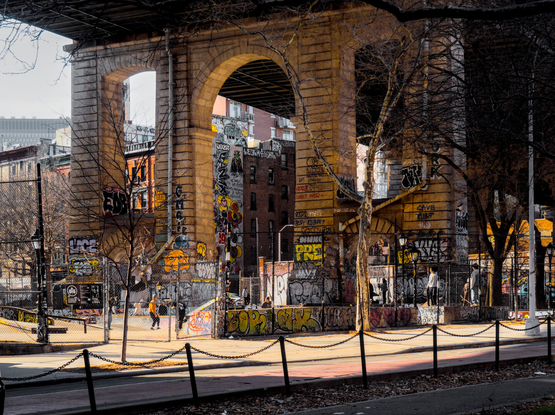 A skate park with lots of graffiti below the giant arches of a bridge. Behind the bridge are old brick mid-rise buildings, also with graffiti.