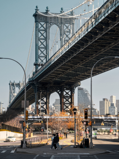 Looking from beneath a great suspension bridge. A bike path curves below the bridge. A few people were walking and biking on the bike path, dwarfed by the bridge.