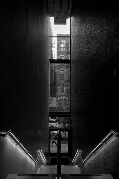 Looking down inside a tall and straight staircase ending at a tall window showing old building outside. The brick walls reflect light from outside. A person was walking by the window.