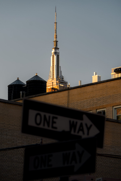The Art Deco spire of Empire State Building shining in the golden afternoon sun rising behind dilapidated residential buildings, one of which has 2 water towers. In the foreground, in the shade and out of focus, is a One Way sign.