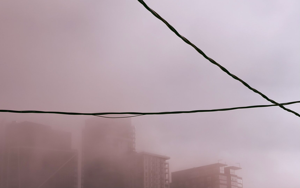 Silhouette of the top floors of high rise buildings, some details visible through low heavy fog. Foreground has 2 crossed overhead wires.