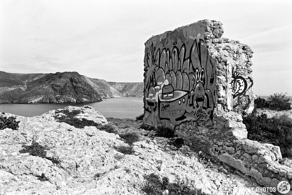 A partially collapsed wall with colorful graffiti sits atop rocky terrain, overlooking a serene body of water and a distant mountainous landscape under a cloudy sky.