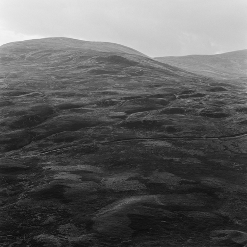 Monochrome photograph of a rolling Highland hillside, its surface textured with peat hags and tussock grass. The hill rises to a broad, rounded summit against a flat grey sky. No human presence, the landscape fills the entire frame.