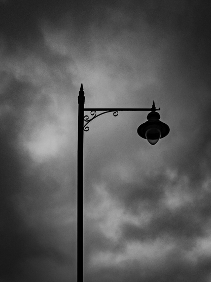 A minimalist black-and-white silhouette of a vintage street lamp positioned against a backdrop of thick, moody clouds.
