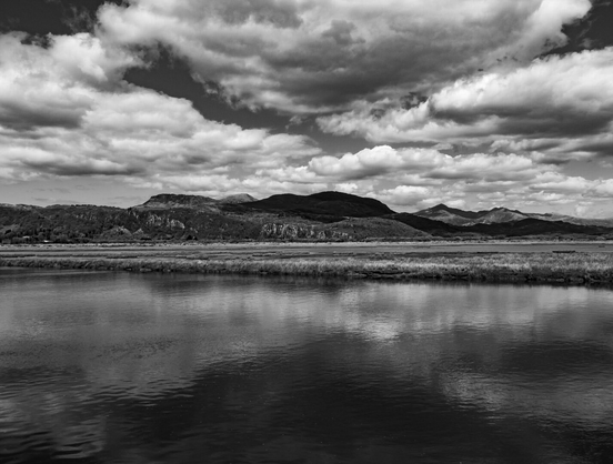 A high-contrast monochrome landscape looking over the Glaslyn estuary toward the dark, prominent peak of Moel Ddu. The dramatic, cloudy sky is reflected in the still water.