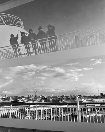 A reflection of a silhouetted group of five people standing on a ship deck railing overlooking an urban waterfront under a cloudy sky.