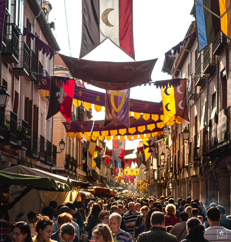 Color photo taken in the late afternoon of a street in an old Spanish town packed full of people with many colorful flags and banners hanging overhead.