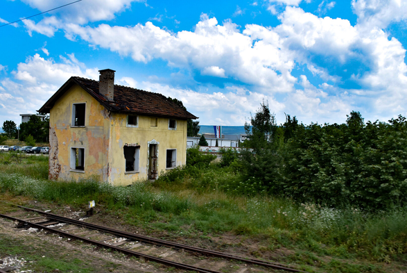 A view from a train window on the route between Istanbul and Sofia, showing a crumbling, abandoned building. The yellow-plastered structure with a red tiled roof is framed by green vegetation and a vibrant blue sky, with a single train track running in the foreground.