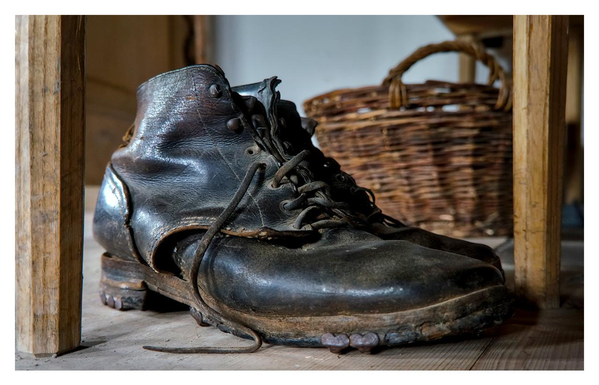 This is a realistic, close-up photograph of two old leather work boots standing one behind the other on a wooden floor. The front boot is fully visible and dominates the image. It is a dark brown to black, high-ankle leather boot with a thick, rugged sole. The leather is heavily worn, cracked, and shiny in places from years of use, especially on the toe cap and sides. The top edge is torn and folded outward, revealing the lighter inner lining. Dark, frayed laces are loosely threaded through the metal eyelets but are untied; one long lace hangs down and trails across the wooden floor in the foreground. The thick heel and front sole show visible metal hobnails or cleats. Directly behind this front boot, a second identical work boot is partially visible, standing in line behind it. Only the upper part of the second boot can be seen, slightly out of focus and tucked between the wooden legs of a piece of furniture. Both boots are positioned between two thick, vertical wooden legs (probably from a table or chair). In the softly lit background, a round woven wicker basket with a curved handle is visible, slightly out of focus. The scene feels rustic, weathered, and nostalgic. Natural light creates gentle highlights on the glossy, worn leather and deep shadows in the cracks and creases of the boots. The overall atmosphere is quiet and old-fashioned, as if these boots have been left behind after many years of hard work.