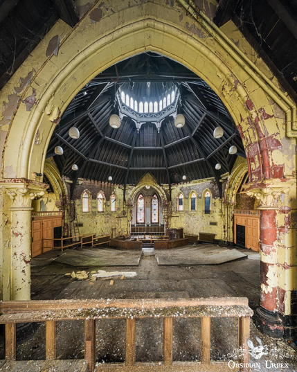 An interior view of an abandoned Victorian chapel, seen through a foreground arch. Peeling yellow paint and exposed brick mark the decaying walls. A central domed skylight illuminates the space.
