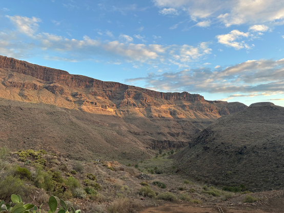 An evening panorama of a mountain valley, with a distant valley wall lit by the setting sun. A road could be seen climbing up the valley,clinging to its side.