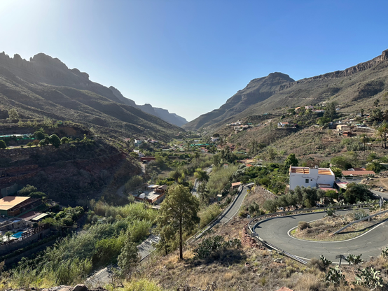 A hamlet of Ayagaures. A group of whitewashed houses is sitting along a winding mountain road, surrounded by small groups of trees at the bottom of a mountain valley.