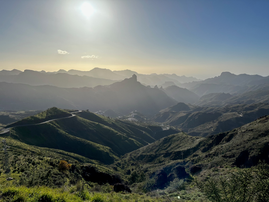 A look down a mountain pass at the start of a new day. The sun is waiting the picture in the hues of yellow. The hills in front are richly overgrown with vegetation. The silhouettes of many mountain ridges are stretching on the horizon.