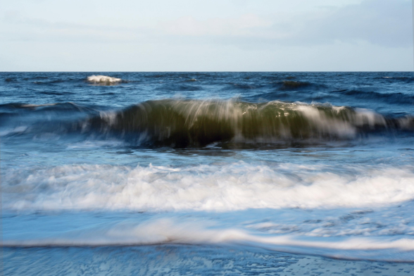 Blick aufs Meer: Wellen schlagen an den - nicht sichtbaren - Strand, die ersten kleineren sind schon gebrochen, die weißen Schaumkronen türmen sich auf. Eine große grünlich-bräunlich wirkende Welle kommt auf das Ufer zu, im Hintergrund ist noch eine weitere kleine Welle mit weißer Krone zu sehen. Der Himmel ist blaugrau. Durch die längere Belichtungszeit wirken Wasser und Wellen wie gemalt.

A view of the sea: waves are crashing onto the – unseen – beach; the first smaller ones have already broken, and the white crests are piling up. A large wave, appearing greenish-brown, is heading towards the shore; in the background, another small wave with a white crest can be seen. The sky is blue-grey. Due to the longer exposure time, the water and waves look as if they have been painted.