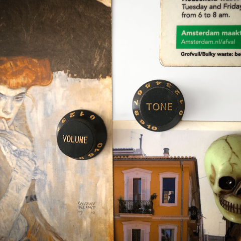 A look at a white bulletin board with a Klimt postcard to the left, a Valencia postard at the bottom, a detial of a green glow in the dark skull magnet at the lower right, part of an Amsterdam PSA magnet at the top right and in the center two round black magnets representing a tone and volume knob on a guitar.