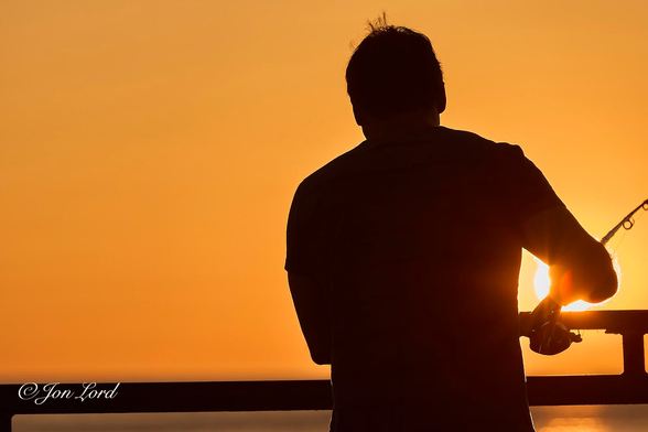 This is a colour photo in landscape format of a man fishing in the sea from a pier at sunset. Redondo Beach, California (2013).

Most of the image is filled with the golden glow of the setting sun. The clear and cloudless sky stretches downwards from the top of the photo to just above the base. Extending across the bottom of the frame is the small amount of sea that's visible below the horizon. Also spreading across the base of the image is the metal railing on the edge of the pier. Our fisherman (Angler) is standing to the right of centre, facing away from the camera and viewed in silhouette against the golden sky. He is holding a fishing rod (fishing pole) in his right hand with his right elbow extended and partially covering the sun, the rim of which can be seen circling his arm. Below the sun's orb and fisherman's arm: the sun's reflection is stretching from the horizon along the Pacific Ocean and towards the camera. 

