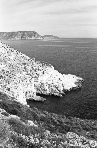 Black and white coastal landscape featuring rugged cliffs meeting the calm sea, with distant hills visible on the horizon. The scene captures the serene beauty of nature, highlighting the textural contrast of the rocky shore and water.