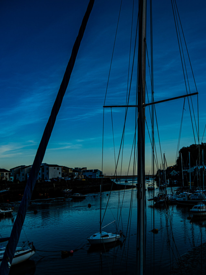 Sailboats docked at Porthmadog Harbour in Wales during twilight. The sky is a dark blue with a hint of orange sunset on the horizon. Silhouettes of yacht masts, and distant village lights reflect on the water.