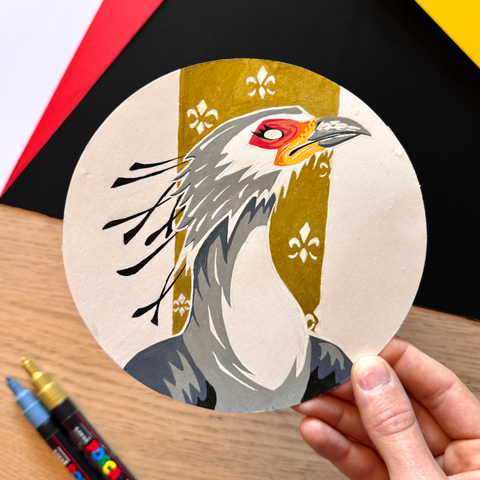 Picture of my hand holding  circle-shaped original illustration painted with poscas featuring a portrait of a secretary bird in front of a gold background decorated with white heraldic lilies. 
