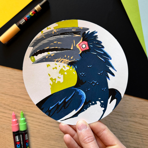 Picture of my hand holding  circle-shaped original illustration painted with poscas featuring a portrait of a hornbill holding a small berry in its beak. 