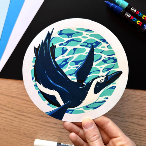 Picture of my hand holding  circle-shaped original illustration painted with poscas featuring a swimming penguin in front of a watery background.