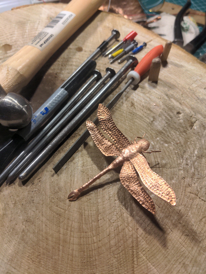 A Copper Dragonfly on a rough wood surface. About 10 cm in size. In the Background is a jewellers hammer, a metal file and a few nails with differently filed tips, that were used as chisels.
