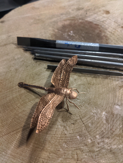 A Copper Dragonfly on a rough wood surface. About 10 cm in size. In the Background is a chisel and a few nails with differently filed tips, that were used as chisels.