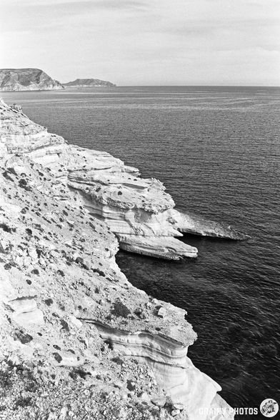 A black and white image capturing a rocky coastline with layered cliffs meeting the calm sea, under a clear sky. In the distance, a small island is visible, adding depth to the scenic view.